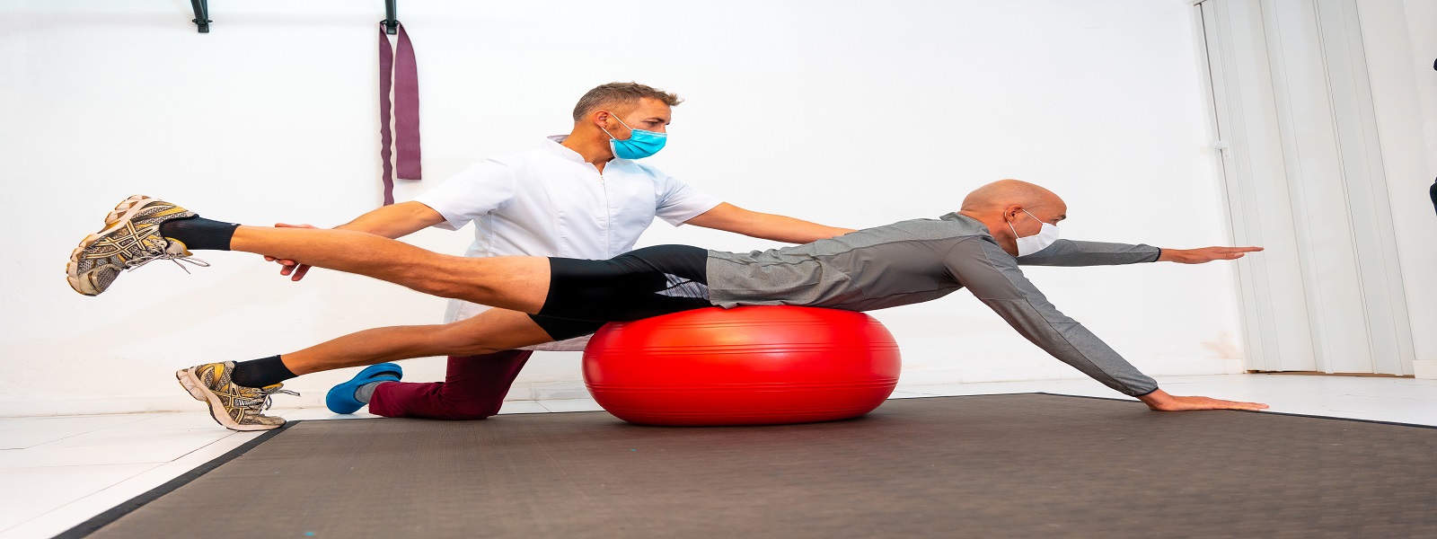 Client balancing on a red ball assisted by Physiotherapist with face mask. Physiotherapy with protective measures for the Coronavirus pandemic, COVID-19. Osteopathy, sports quiromassage